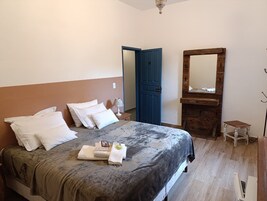 A neatly made bed with white pillows and a grey bedspread, a wooden dresser with a mirror, and a blue door in the background.
