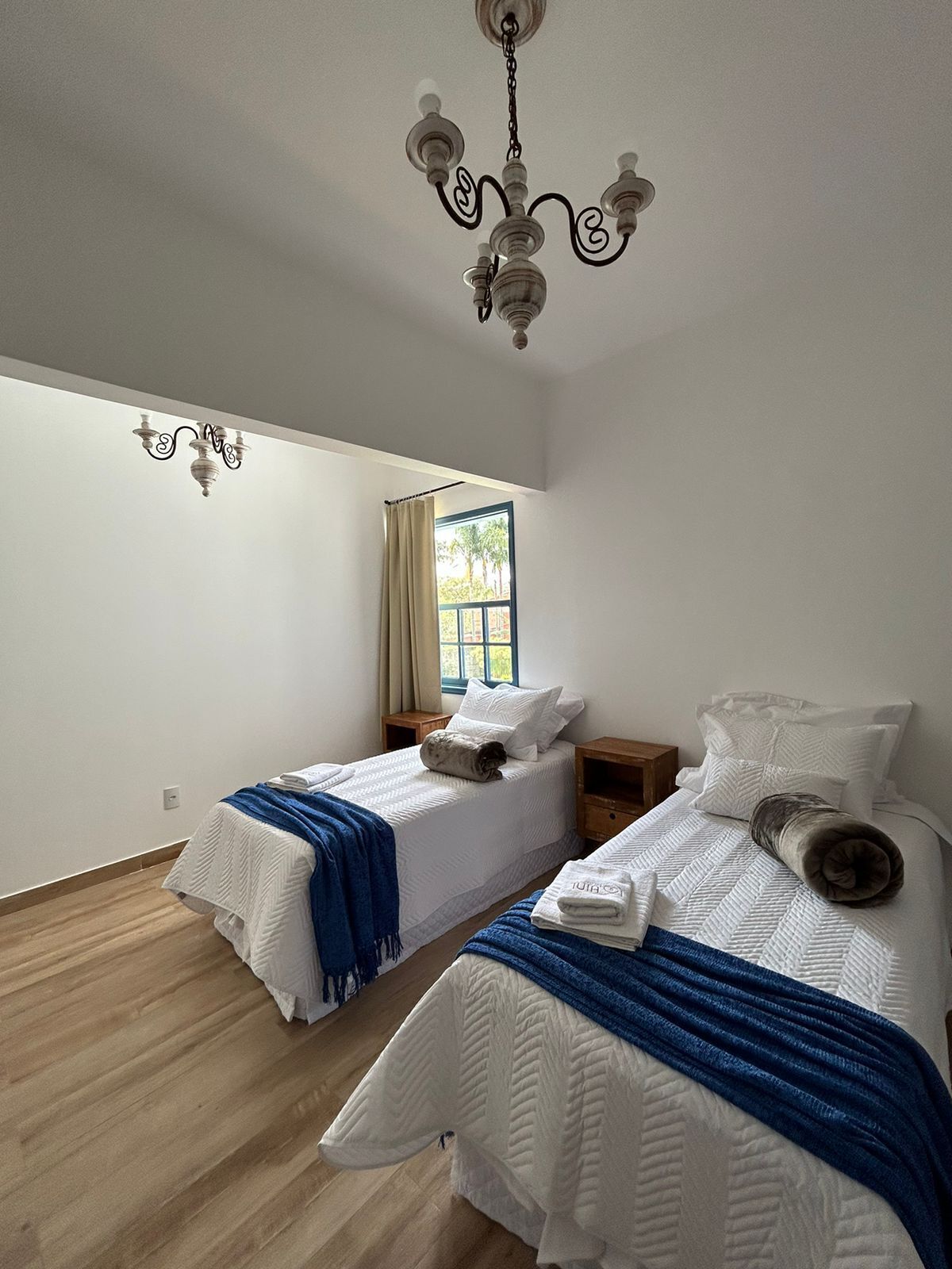 A bedroom with two beds, a chandelier, a window with curtains, and a wooden floor.