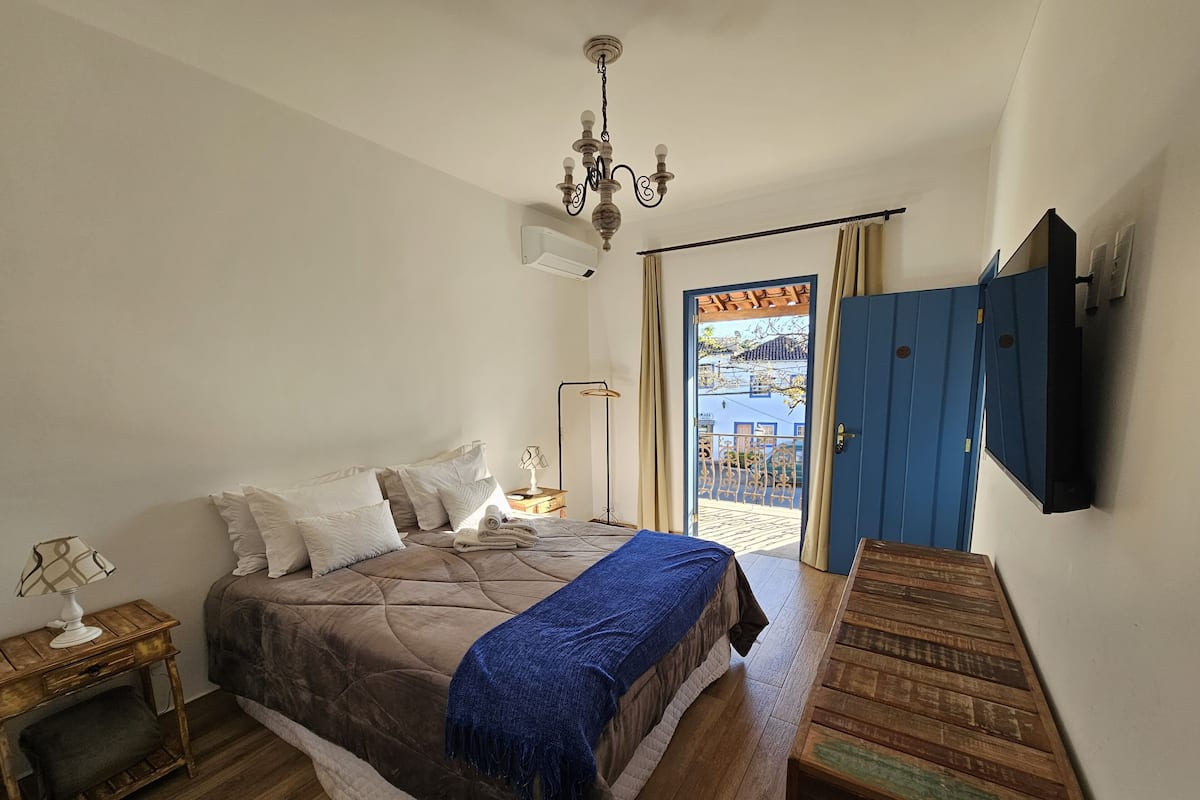 A bedroom with a bed, bedside table, lamp, and a view of the outside.