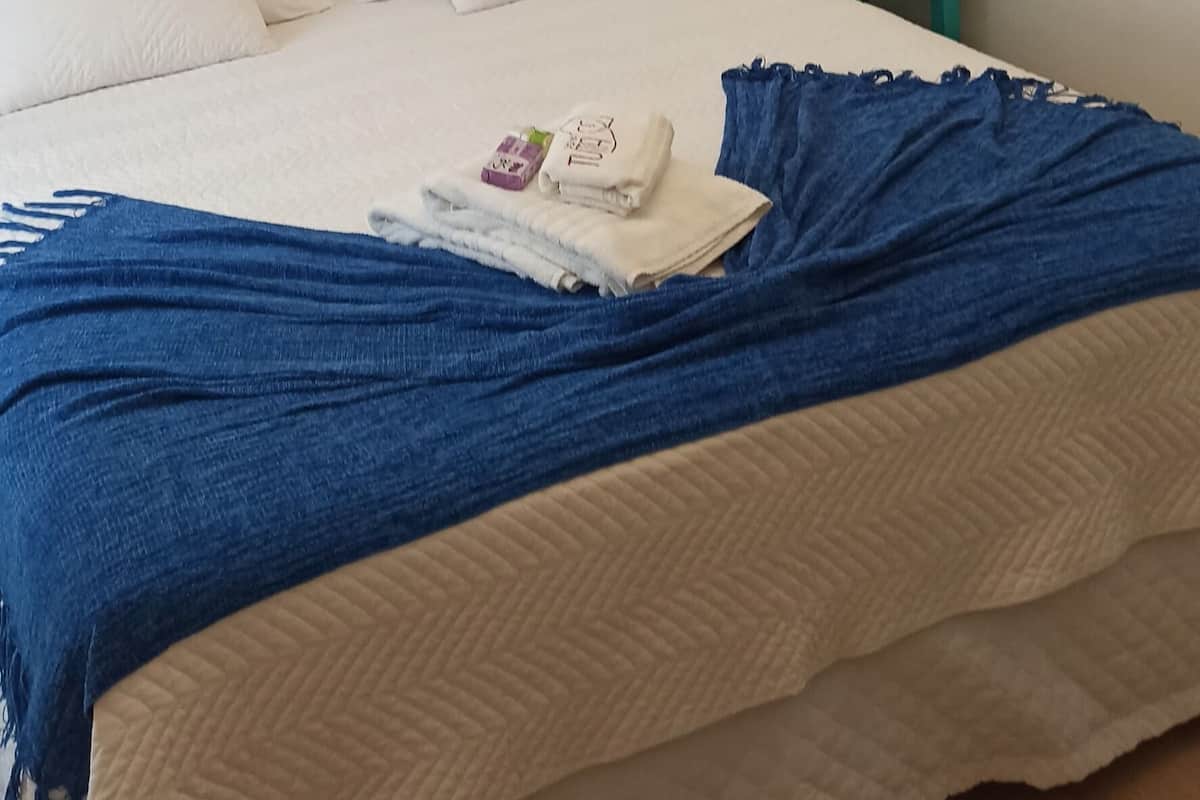 A neatly made bed with white linens and a blue blanket, a bedside table with a lamp, and a wooden floor.