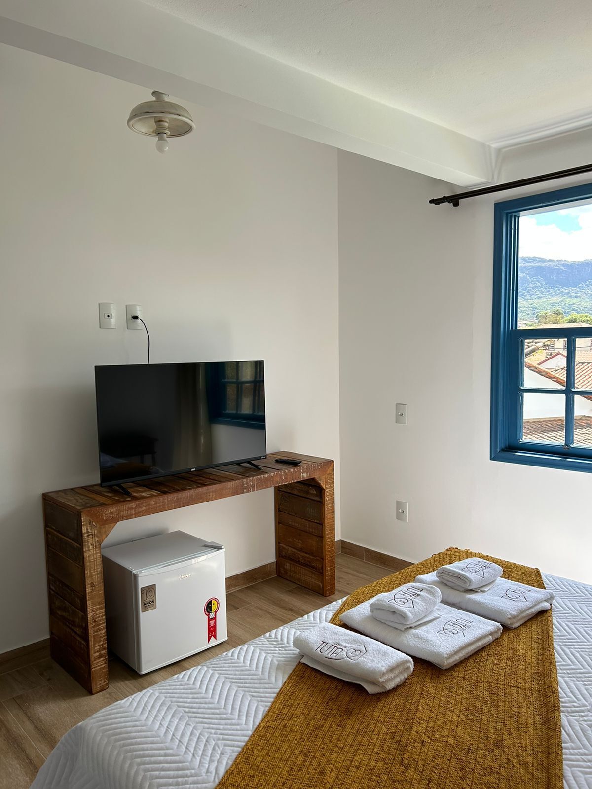A bedroom with a bed, a wooden desk with a TV, a mini-fridge, and a window with a view of the outdoors.