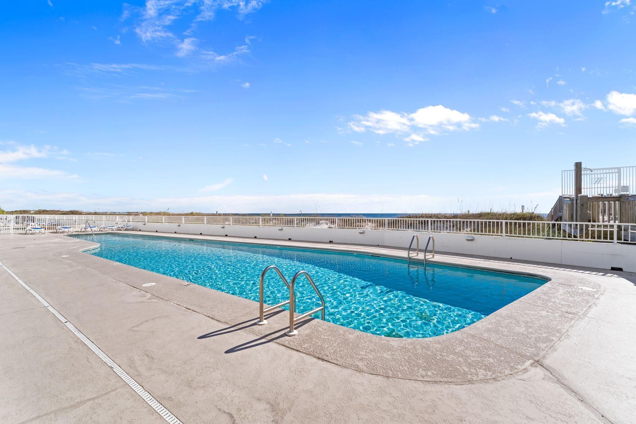 Une piscine rectangulaire à l’eau d’un bleu cristallin, entourée d’une terrasse blanche et de rampes en métal.