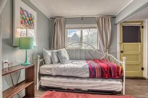 A bedroom with a bed, a bedside table, a lamp, a window with curtains, and a door. - Enchanted Garden Studio: Cozy Downtown (Fruita)