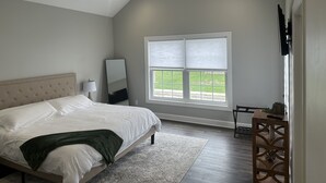 A bedroom with a bed, a ceiling fan, a large window with blinds, and a small table with a remote control. - NEWLY BUILT condo - premium features, game room, firepit, short walk to village (Ellicottville)