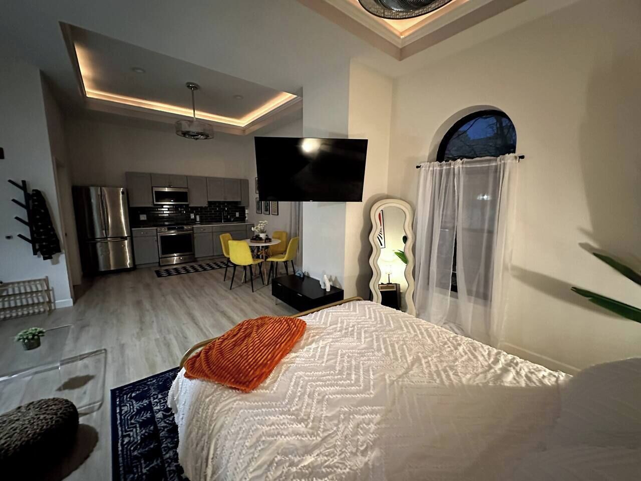 A modern hotel room with a bed, a TV, a kitchen area, and a dining space.