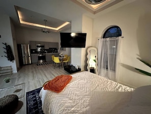 A modern hotel room with a bed, a TV, a kitchen area, and a dining space.