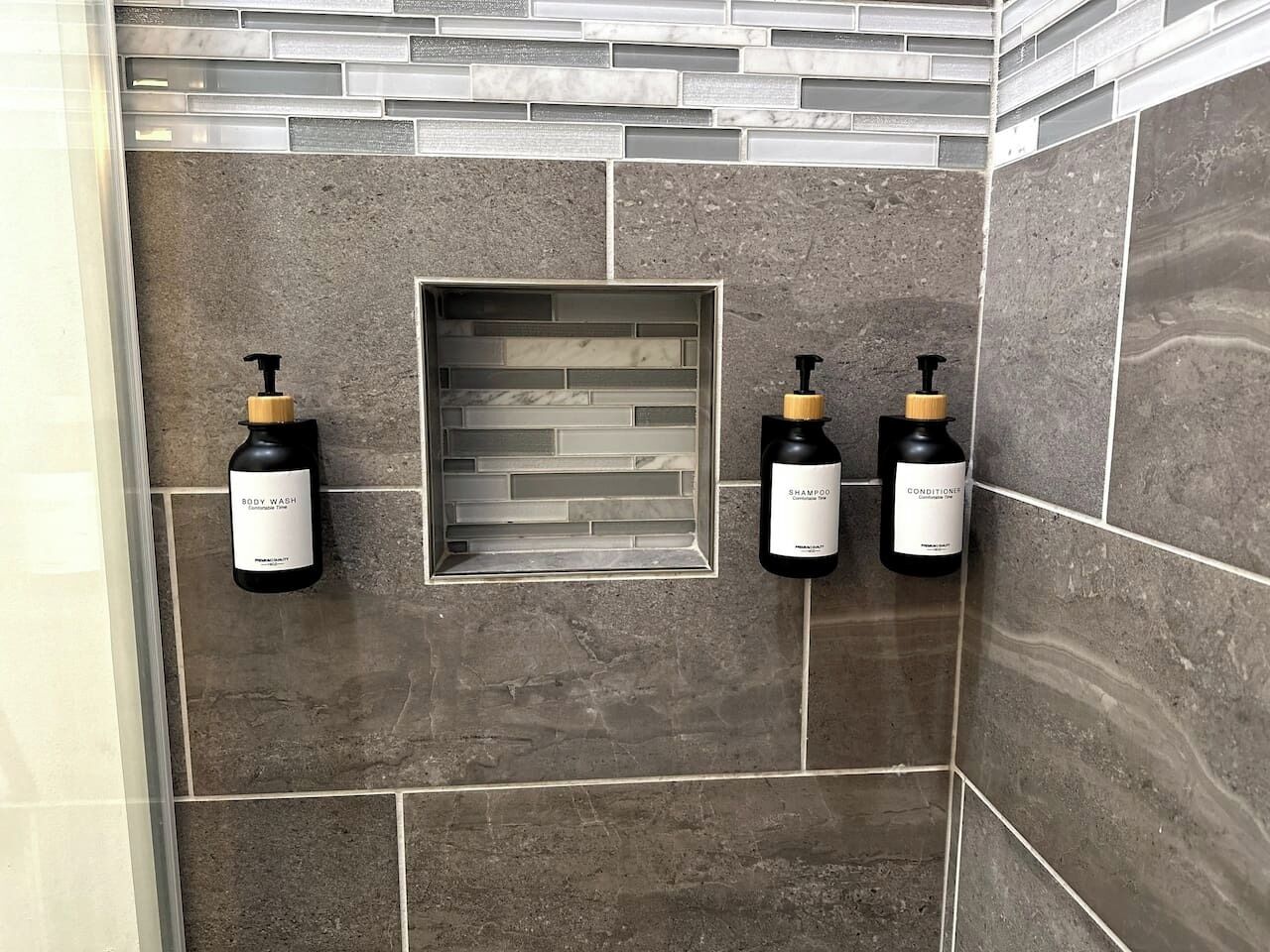 A bathroom with a tiled wall, a built-in shelf, and three bottles of shampoo and conditioner.