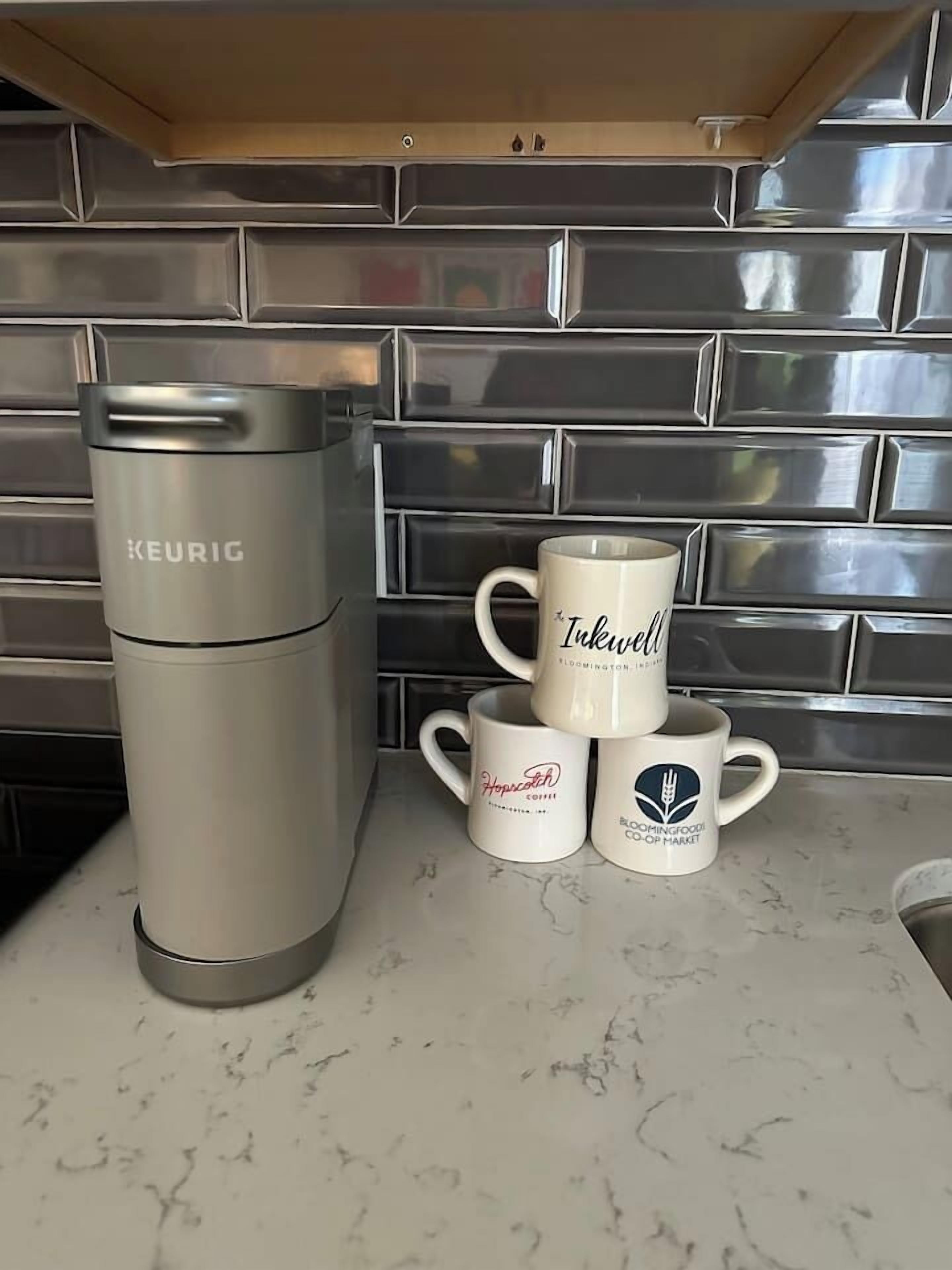 En Keurig-kaffemaskin, två muggar med logotyperna för ”Iwakwell” och ”Bloomingfood Co-op Market”, och en stapel med tre muggar på en bänkskiva i marmor.