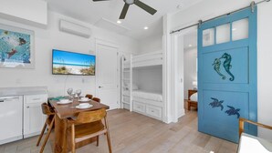 A modern kitchen with white cabinets, a dining table set for four, and a large flat-screen TV mounted on the wall.