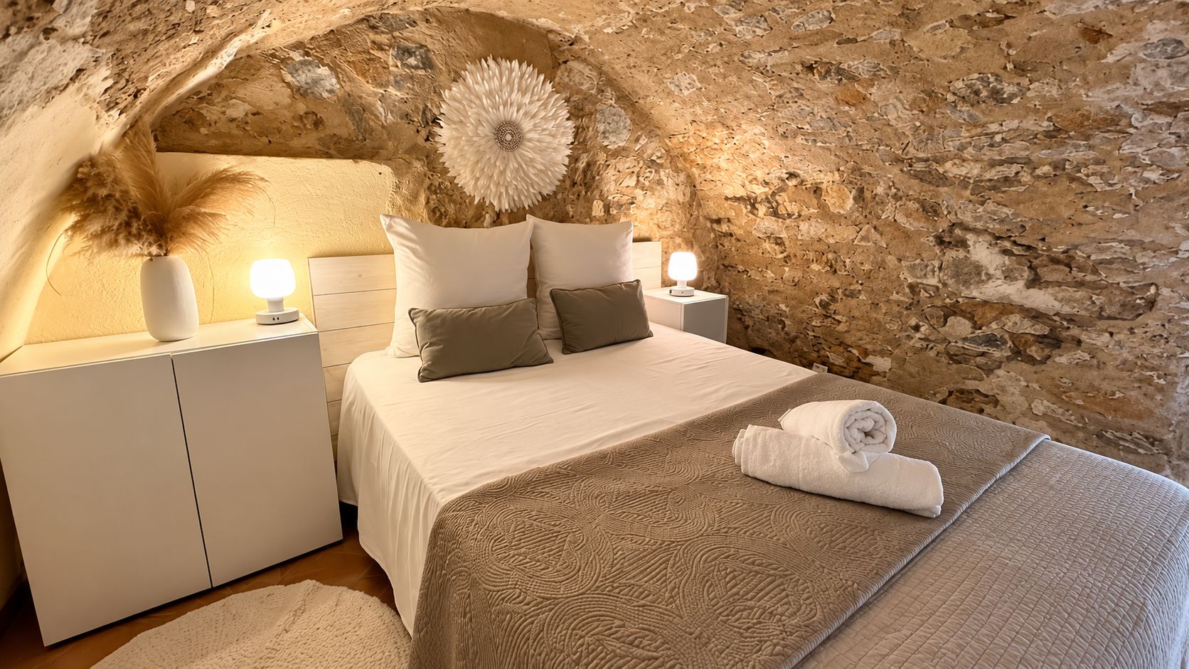 Une chambre avec un mur en pierre, un lit recouvert d’une couvre-lit à motifs, deux tables de chevet avec des lampes et un élément décoratif mural.