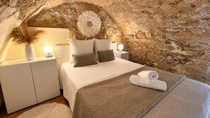 A bedroom with a stone wall, a bed with a patterned bedspread, two bedside tables with lamps, and a decorative wall piece.