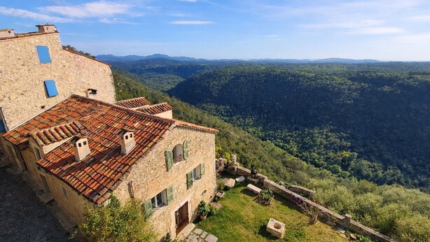 A stone building with a tiled roof, green shutters, and a view of a forested valley.