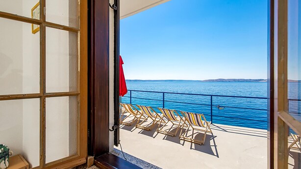En åben dør, der fører ud til en balkon med havudsigt og en række liggestole.