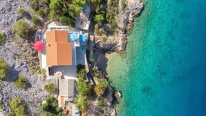 Oversigtsbillede af en kystejendom med et hus, en swimmingpool og krystalklart turkis vand.