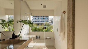 Un baño moderno con un espejo grande, un tocador de madera, cabina de ducha de vidrio y vistas a un edificio con balcones.