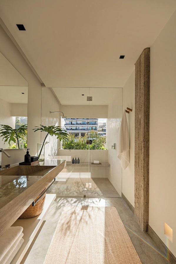 Une salle de bain moderne dotée d’un grand miroir, d’un meuble-lavabo en bois, d’une cabine de douche en verre et offrant une vue sur un immeuble avec des balcons.