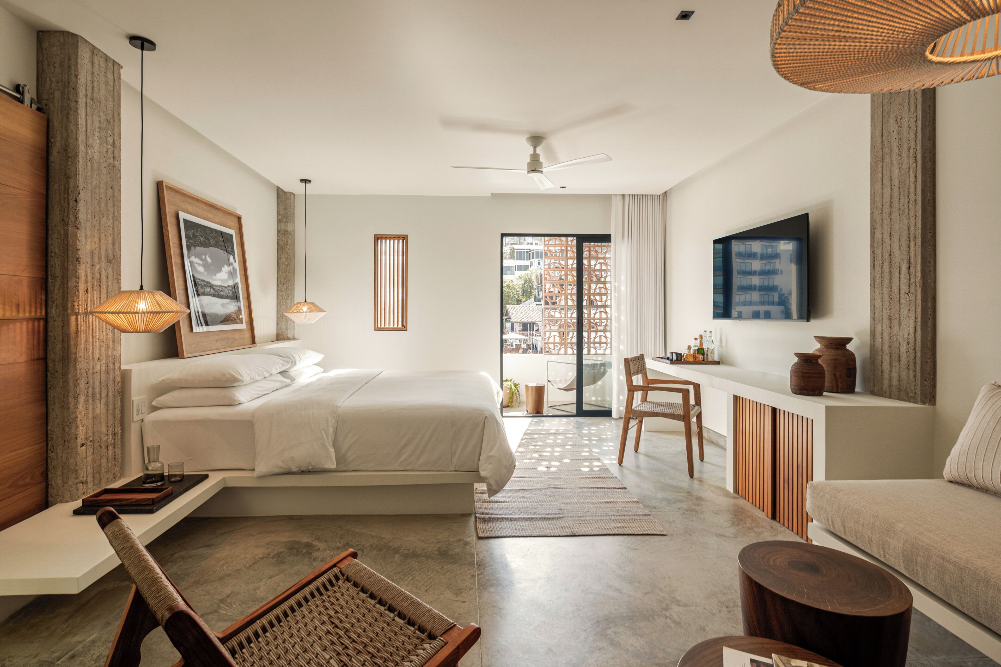 Une chambre d’hôtel moderne, dotée d’un grand lit, d’un bureau, d’une chaise et d’un canapé. Un téléviseur est fixé au mur et une fenêtre offre une vue sur les bâtiments environnants.