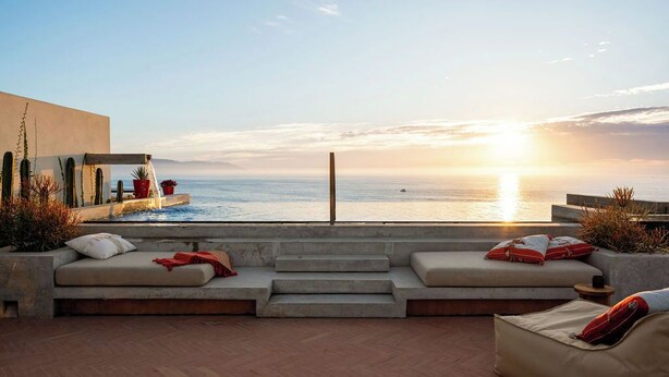 Una terraza con vistas a la puesta de sol sobre el mar, con un espacio de descanso con cojines y una pequeña mesa.