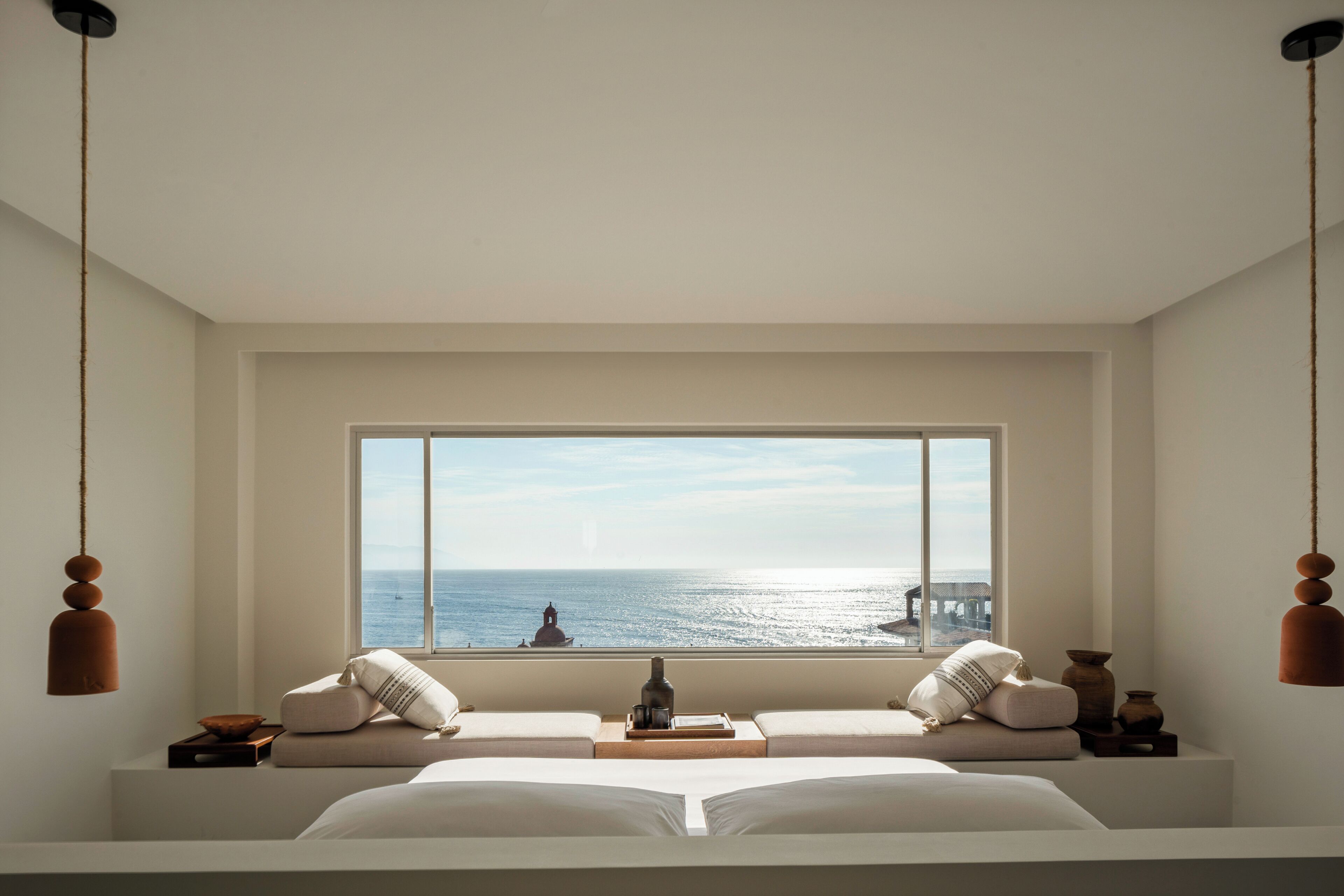Une chambre avec une grande fenêtre donnant sur la mer, un lit, une table de chevet et des lampes suspendues.
