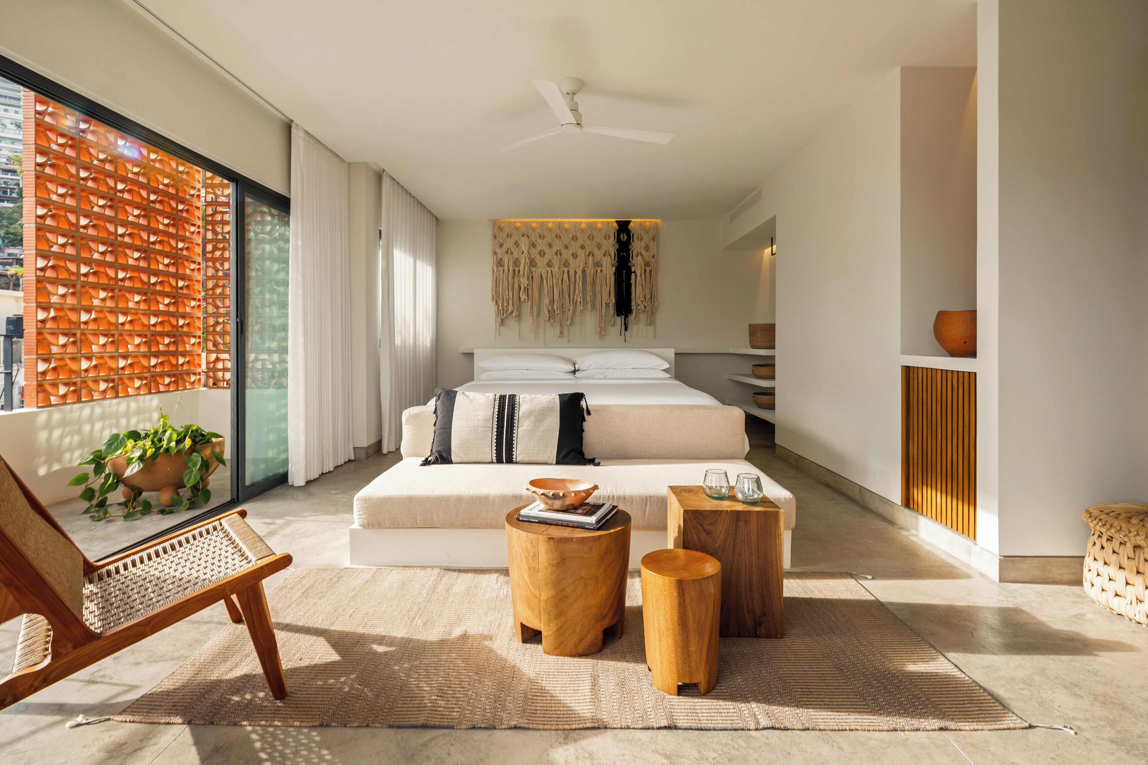 Une chambre moderne avec un grand lit, un coin salon comprenant un fauteuil et des tables d’appoint, et une vue dégagée sur l’extérieur grâce à une grande fenêtre.