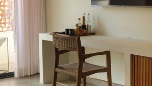 Una sala de estar moderna con un televisor de pantalla plana grande montado en la pared, una silla de madera, una mesita blanca con una bandeja de objetos y un ventanal con cortinas.