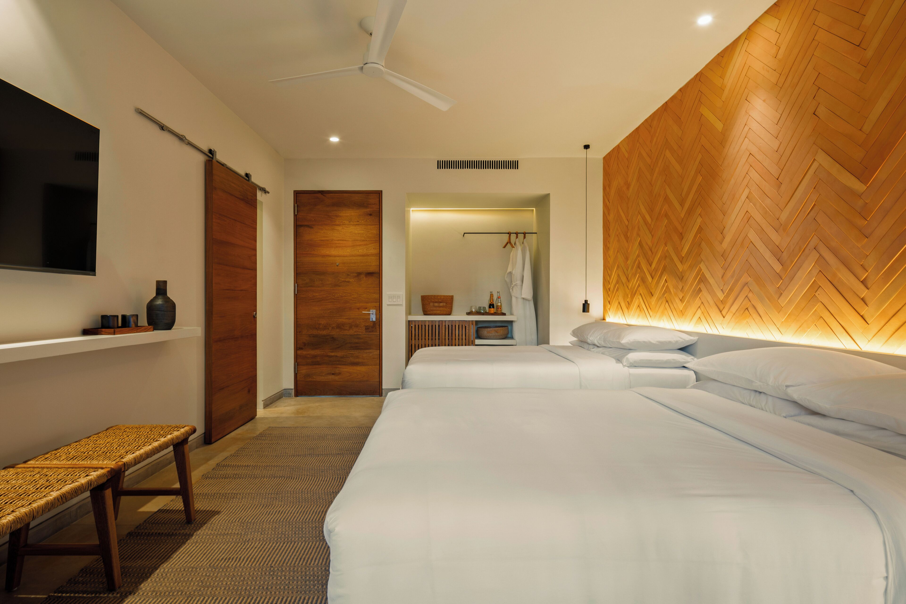 Une chambre d’hôtel moderne avec deux lits, un banc, une télévision et un mur recouvert de lambris en bois disposés en motif d’arête de poisson.