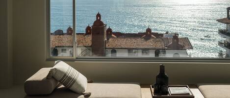 A room with a large window overlooking a coastal view, a sofa with cushions, and a bottle on a tray.