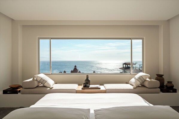A bedroom with a large window overlooking the sea, a bed with white linens, and a wooden nightstand with a bottle and glasses. - The Tryst Puerto Vallarta LGBT Hotel (Puerto Vallarta)