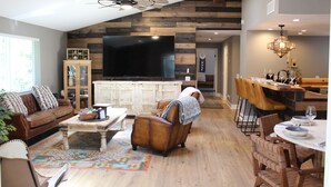 A spacious living room with a large flat-screen TV, a wooden ceiling, and a dining area with a bar. - Luxury Mountain Home with Fireplace, Yosemite & Bass Lake (Oakhurst)
