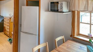 Une cuisine compacte avec un réfrigérateur blanc, un four à micro-ondes posé sur une étagère en bois, une table à manger en bois avec des chaises, et une fenêtre donnant sur des arbres.