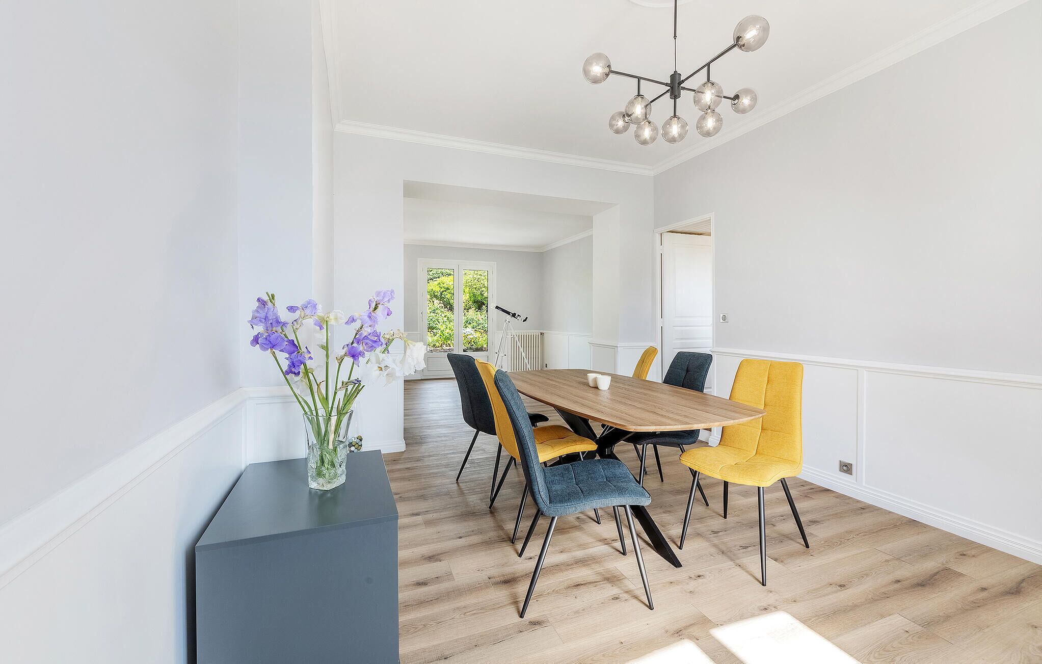 Uma sala de jantar com mesa de madeira, cadeiras coloridas e um lustre moderno.