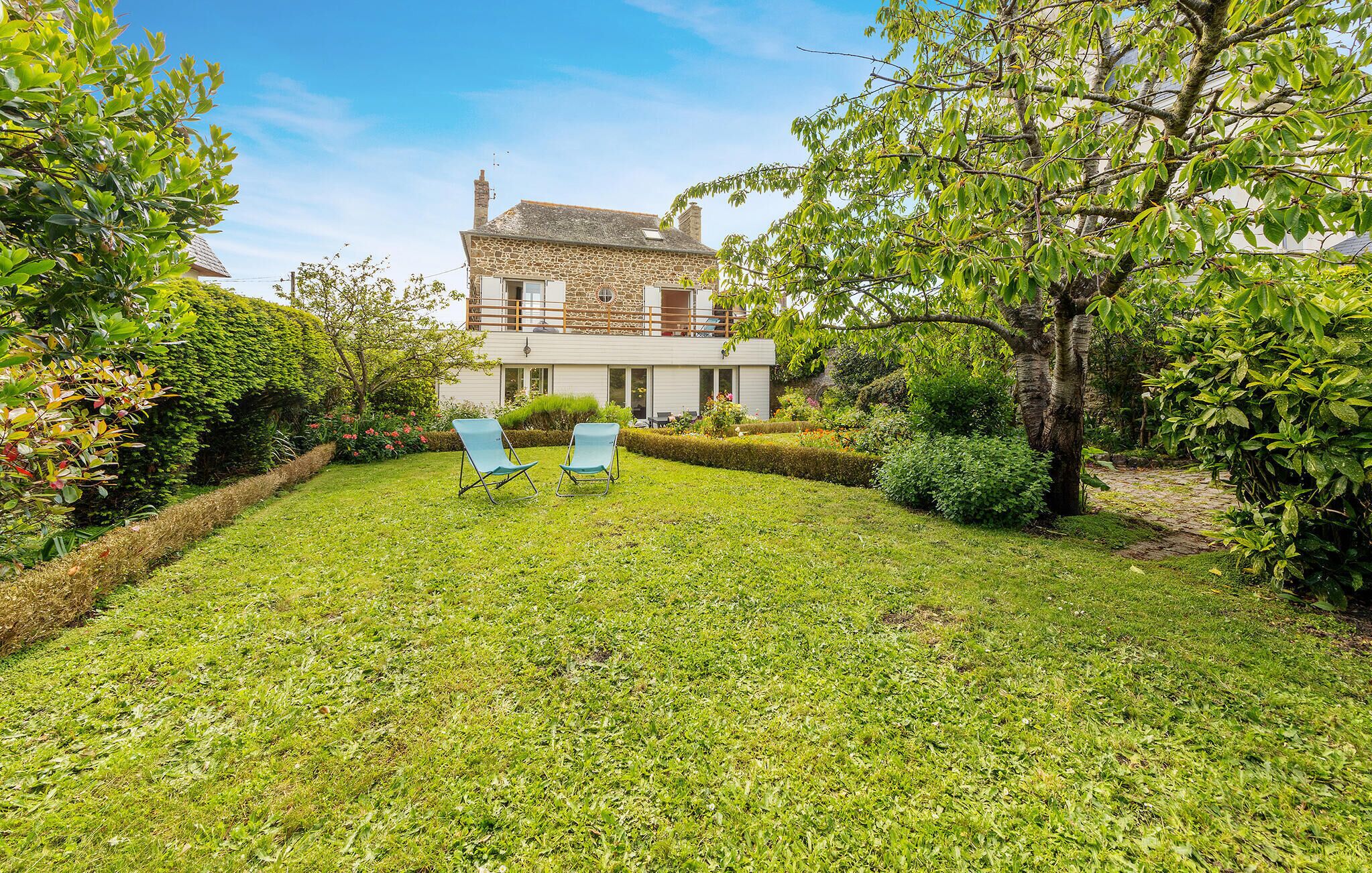 A stone house with a balcony, surrounded by a well-maintained garden and two blue chairs.