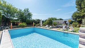 Una piscina rectangular con agua azul cristalina, rodeada de sillas de descanso y sombrillas, con árboles y un cielo despejado al fondo.