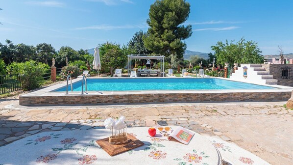 Una piscina con un pérgola, baldosas de piedra y una mesa con un periódico, flores y una pelota roja.
