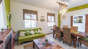 A living room with a green sofa, wooden dining table, wicker chairs, and a hanging light fixture. - Villa Cuqui Antequera by Ruralidays (Málaga)