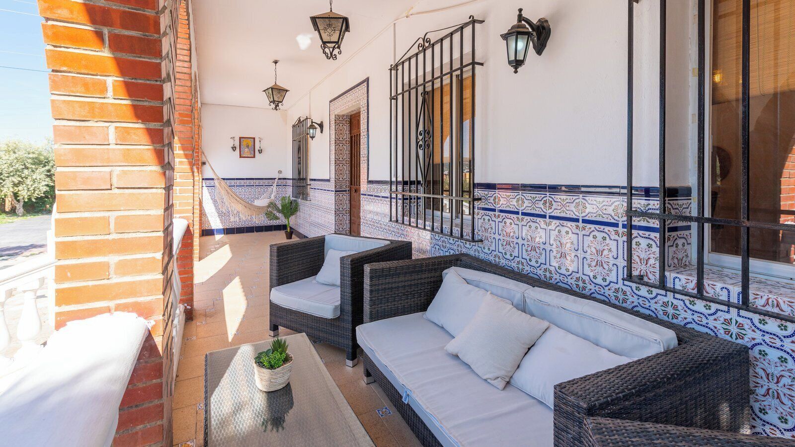 A balcony with wicker furniture, a potted plant, and a tiled wall with a blue and white pattern.