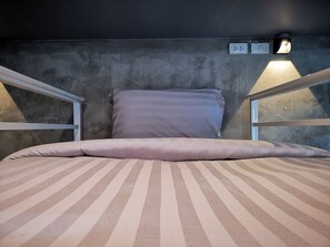 A bunk bed with a striped mattress and pillows, a concrete wall, and a wall-mounted light fixture.
