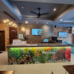 A reception area with a modern design, featuring a 'Welcome' sign, a digital screen displaying breakfast options, and a ceiling fan. - Hi Samui Hostel (Koh Samui)