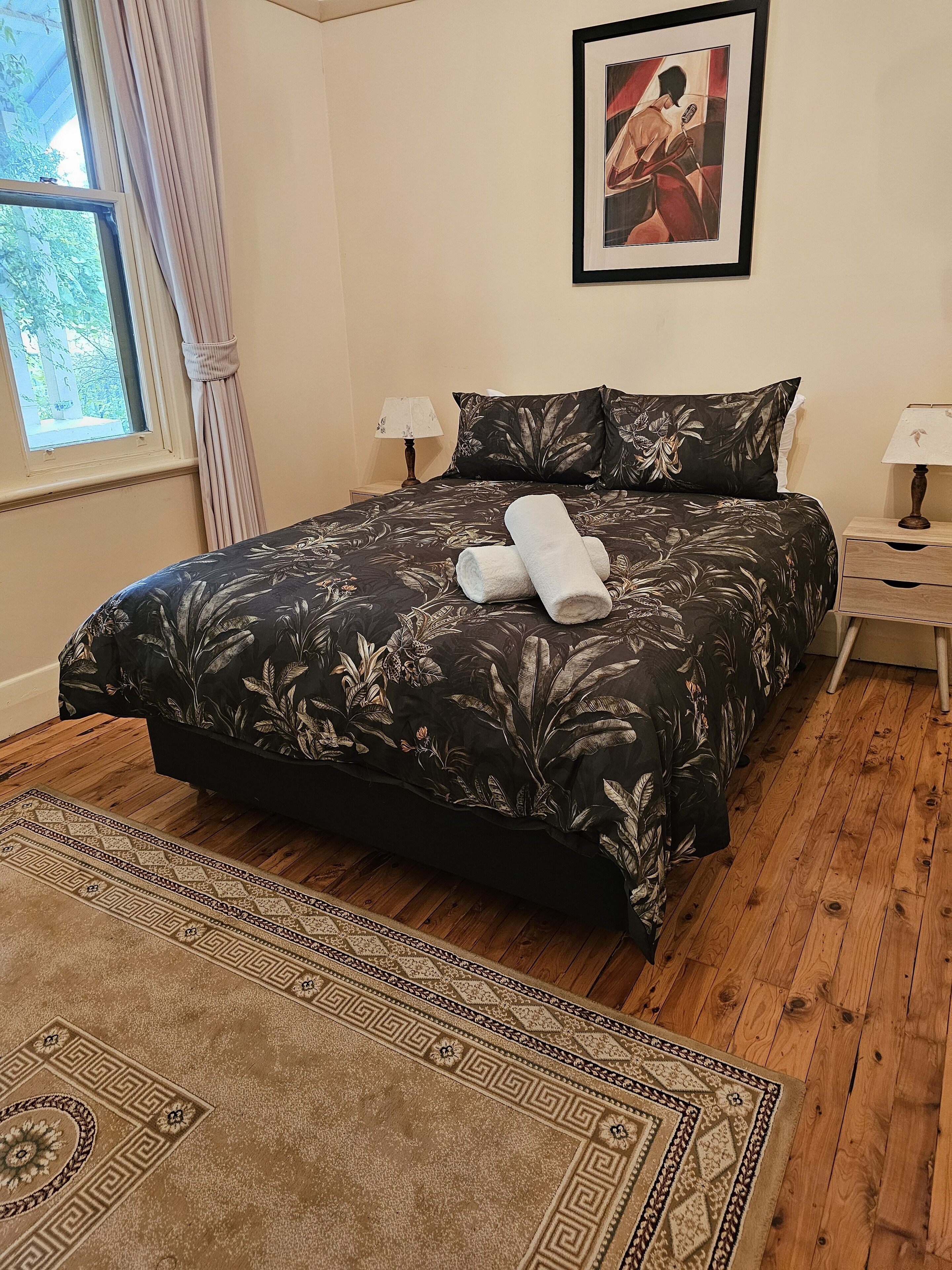 A neatly made bed with a floral bedspread, a pair of rolled towels, a framed picture on the wall, and a wooden floor.