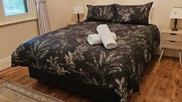 A neatly made bed with a floral bedspread, a pair of rolled towels, a framed picture on the wall, and a wooden floor.
