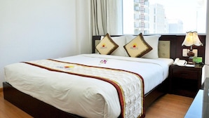 Habitación de hotel con cama, mesita de noche con lámpara, ventana con vistas a edificios y cortina.