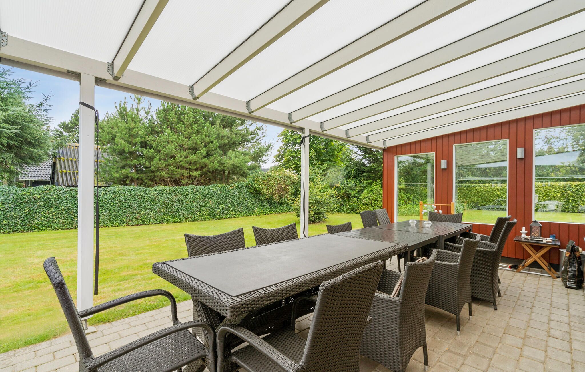 Un patio couvert avec une table et des chaises, donnant sur un jardin, et un bâtiment rouge en arrière-plan.