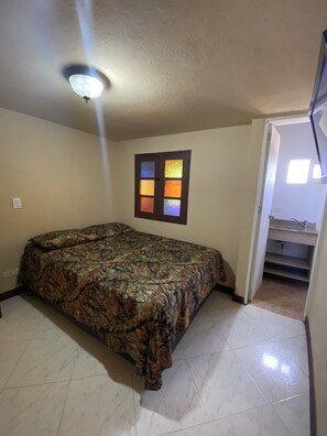 Une chambre à coucher avec un lit, une fenêtre ornée de vitraux, un ventilateur de plafond et une salle de bain visible à travers une porte ouverte.