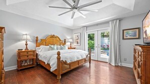 A bedroom with a wooden bed, a ceiling fan, a nightstand with a lamp, a dresser, and a window with a view of greenery. - Pool House 1/2 Mile From Downtown Stuart (Stuart)