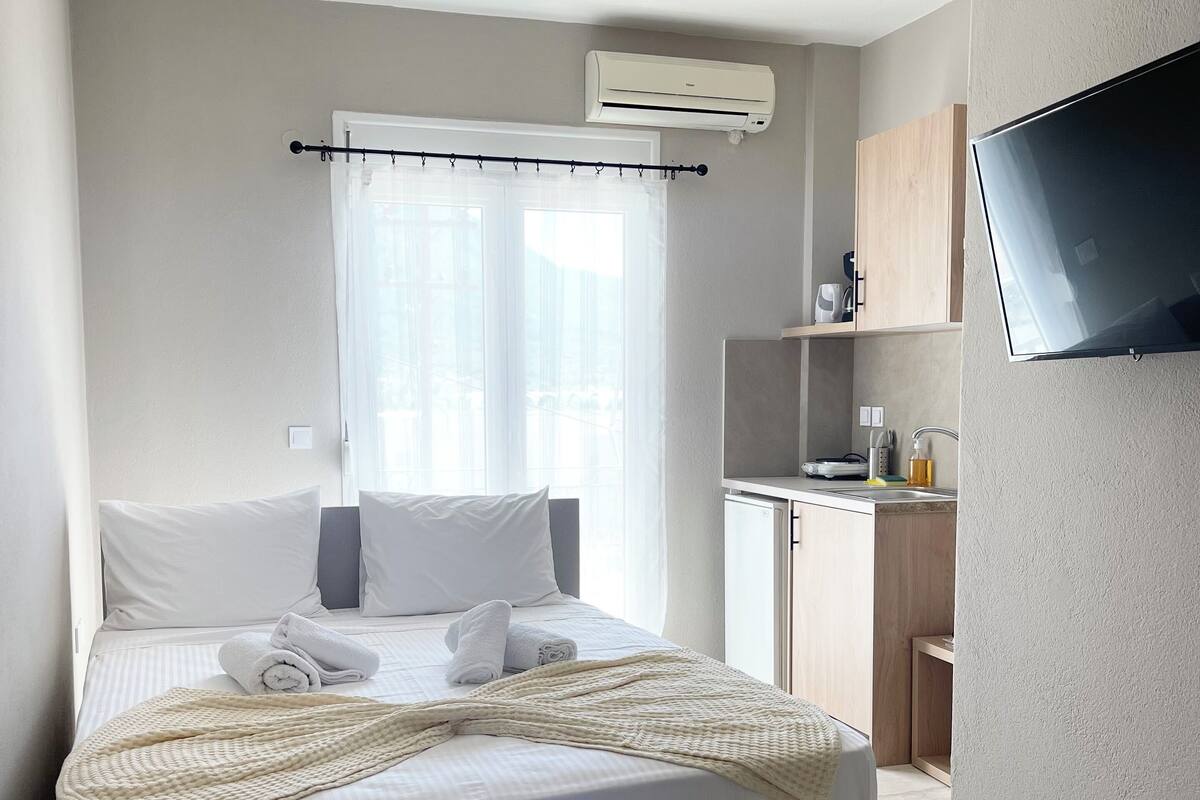 A modern hotel room with a bed, a flat-screen TV mounted on the wall, a small kitchenette, and a window with sheer curtains.