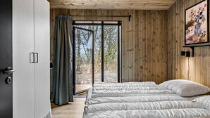 A bedroom with a large bed, wooden walls, a sliding glass door leading to a garden, and a painting on the wall.