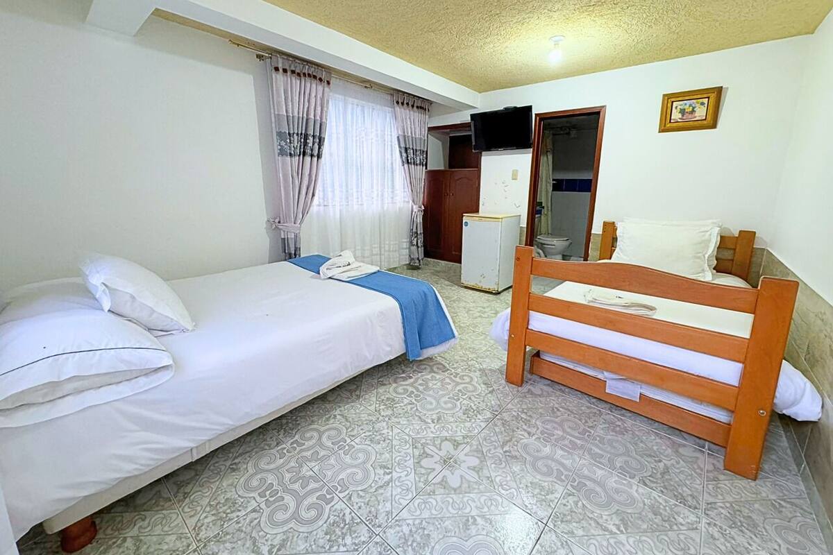 Una habitación pequeña y limpia con dos camas, un banco de madera, un televisor y un baño visible a través de una puerta abierta.