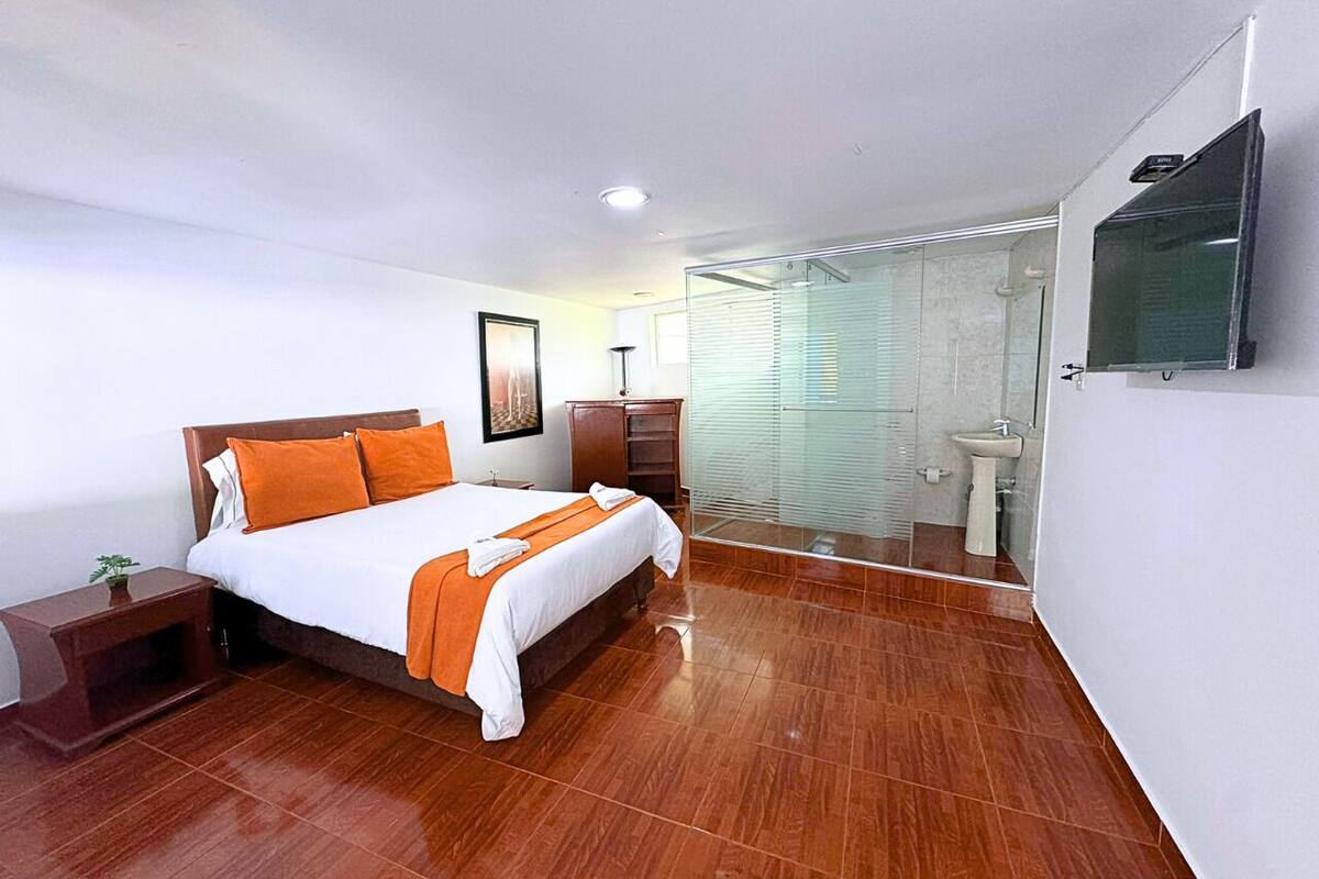Una habitación de hotel moderna con suelo de madera, una cama grande con ropa de cama naranja, una mesita de noche y un televisor montado en la pared, y un baño con cabina de ducha de vidrio.