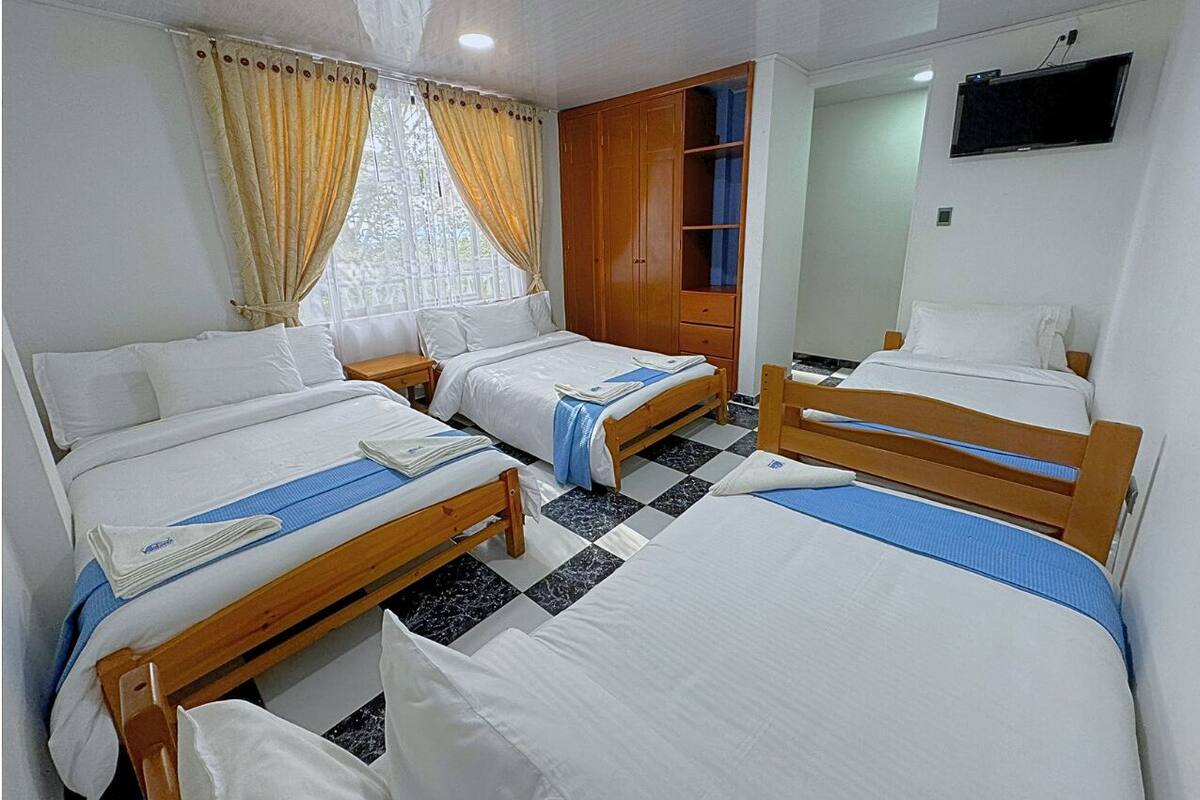 Habitación de hotel con tres camas individuales, un armario de madera, un televisor en la pared y una ventana con cortinas.