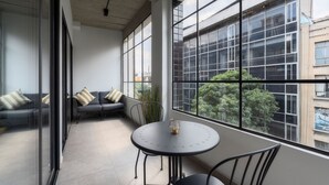 Un balcón con una mesa redonda y sillas, un sofá con cojines, y ventanas amplias que ofrecen vistas de la ciudad.